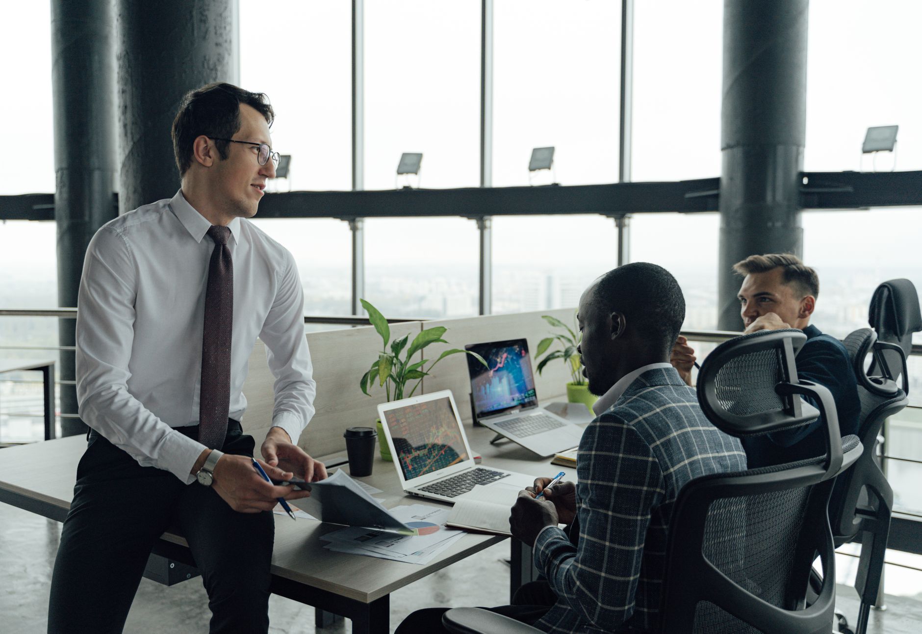Three men engaged in a business meeting in a modern office setting with laptops and documents. — The effects of the current e